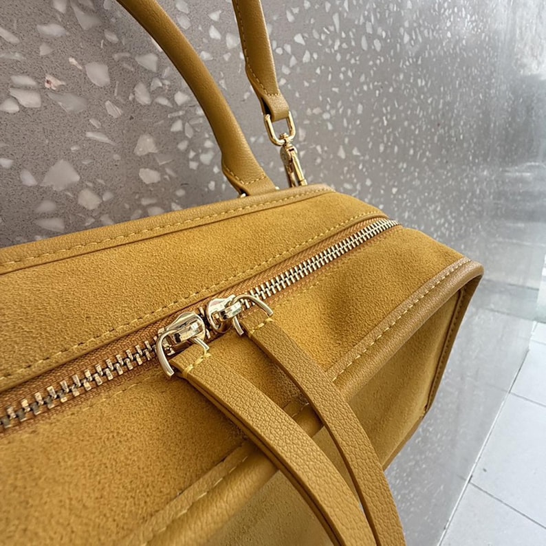 K&ouml;nnte beinhalten: Eine senfgelbe Handtasche aus einem wildlederartigen Material mit einem Rei&szlig;verschluss oben, zwei passenden Zuglaschen, einer strukturierten rechteckigen Form und einem einzelnen Griff, der mit goldfarbenen Metallclips befestigt ist.