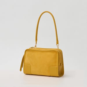 K&ouml;nnte beinhalten: Eine senfgelbe, rechteckige Handtasche mit einer weichen, wildlederartigen Textur am Hauptk&ouml;rper, glatten, lederartigen Eckakzenten, einem einzelnen Schulterriemen, einem Rei&szlig;verschluss an der Oberseite und einer dekorativen Quaste.
