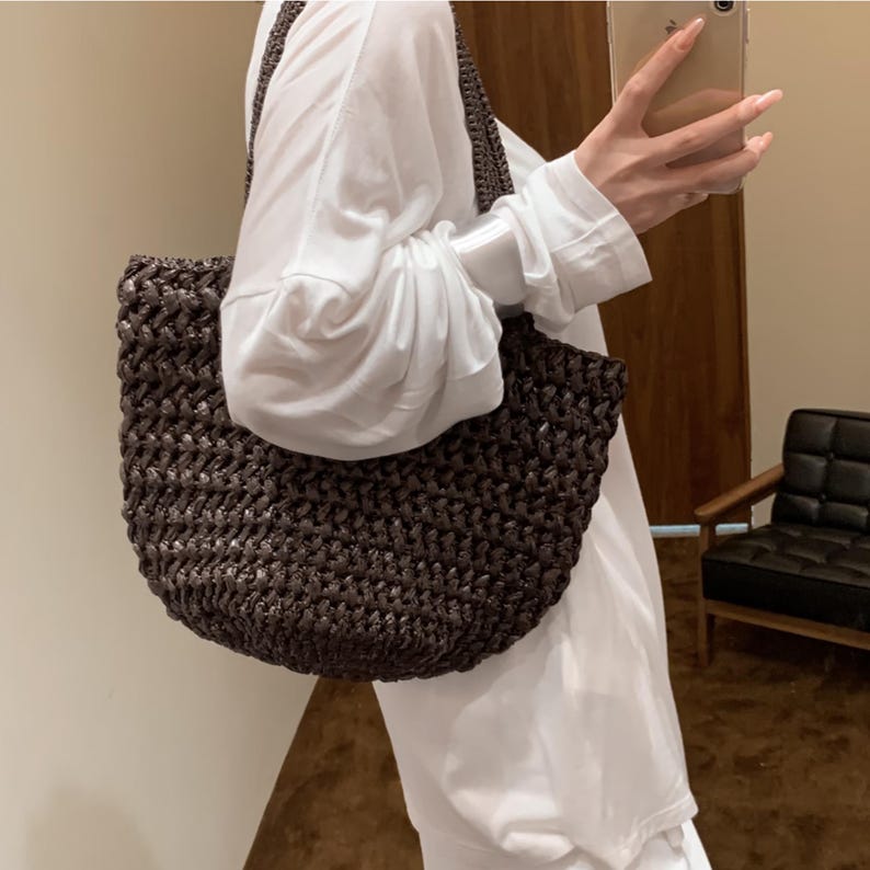 May include: A dark brown woven tote bag with a rounded bottom and a shoulder strap. The bag is made of a textured material and is being held by a person wearing a white long-sleeved shirt.