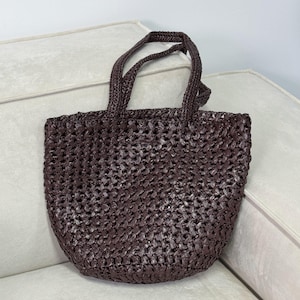 May include: A woven brown tote bag with two handles. The bag has a textured, open weave design and a rounded bottom. The handles are also woven and appear sturdy. The bag is sitting on a light-colored surface.