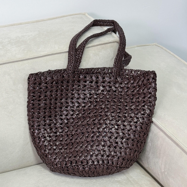 May include: A woven brown tote bag with two handles. The bag has a textured, open weave design and a rounded bottom. The handles are also woven and appear sturdy. The bag is sitting on a light-colored surface.