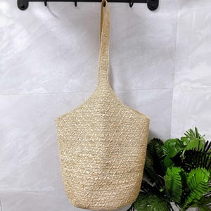 May include: A beige woven tote bag with a long handle, hanging against a light gray tiled wall. The bag has a diamond pattern and a wide opening. The handle is made of the same woven material. The bag is empty.