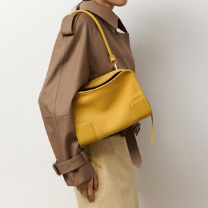 K&ouml;nnte beinhalten: Eine leuchtend gelbe Lederschultertasche mit rechteckiger Silhouette, glatter Oberfl&auml;che, einem einzelnen Schulterriemen und einem strukturierten Design mit einem markanten quadratischen Aufn&auml;her an der unteren Ecke der Vorderseite.