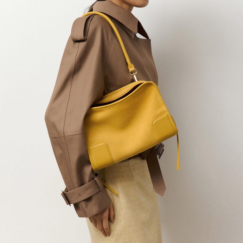 K&ouml;nnte beinhalten: Eine leuchtend gelbe Lederschultertasche mit rechteckiger Silhouette, glatter Oberfl&auml;che, einem einzelnen Schulterriemen und einem strukturierten Design mit einem markanten quadratischen Aufn&auml;her an der unteren Ecke der Vorderseite.