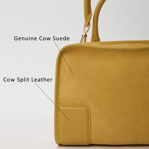 K&ouml;nnte beinhalten: Eine senfgelbe Handtasche mit einer strukturierten Oberfl&auml;che, die als Genuine Cow Suede beschriftet ist, und einem glatten rechteckigen Aufn&auml;her mit der Aufschrift Cow Split Leather. Die Tasche hat einen Tragegriff mit goldfarbenen Beschl&auml;gen.