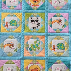 Könnte beinhalten: Eine blaue und gelbe Babydecke mit verschiedenen Tier- und Schmetterlingsmotiven. Die Decke besteht aus Patchwork-Quadraten und hat einen hellblauen Rand. Ein passendes Kissen mit einem Schmetterlingsmotiv ist im Lieferumfang enthalten.