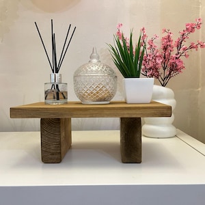 May include: A wooden tiered stand with two levels, featuring a clear glass jar with black diffuser sticks, a clear glass jar with a diamond pattern, and a white ceramic planter with a green plant and pink flowers.