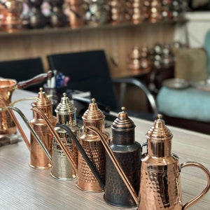 May include: Five copper oil and vinegar cruets with hammered finishes. The cruets have a spout and a handle. The cruets are arranged in a row on a wooden surface.