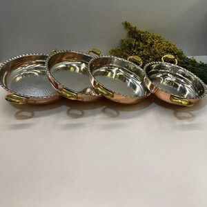 May include: Set of four copper serving bowls with silver interiors and brass handles. The bowls are nested together, with the largest bowl at the bottom and the smallest bowl at the top.