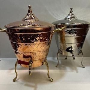 Op de afbeelding: Twee sierlijke metalen dranken dispensers met deksels en tuiten. De dispenser aan de linkerkant is koper met een ingewikkeld bloemmotief en een rode handgreep. De dispenser aan de rechterkant is zilver met een gehamerde afwerking.