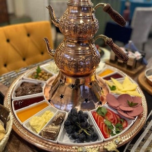 以下が含まれることがあります： 装飾的なデザインが施された銅製のティーセットが、果物、チーズ、スライスした肉など、さまざまな軽食を載せた丸い銅製のトレイの上に置かれています。