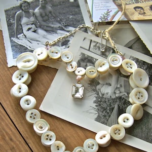 Könnte beinhalten: Eine herzförmige Halskette aus weißen perlmuttartigen Knöpfen mit einem Kristallanhänger und einem kleineren Kristall-Anhänger. Die Halskette wird auf einer Holzoberfläche mit Vintage-Fotos präsentiert.