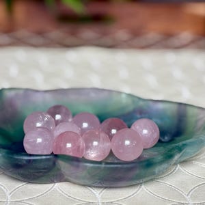 May include: A collection of pink rose quartz spheres displayed in a leaf-shaped dish. The dish has a marbled appearance with shades of green, blue, and purple. The spheres are smooth and translucent, reflecting light.