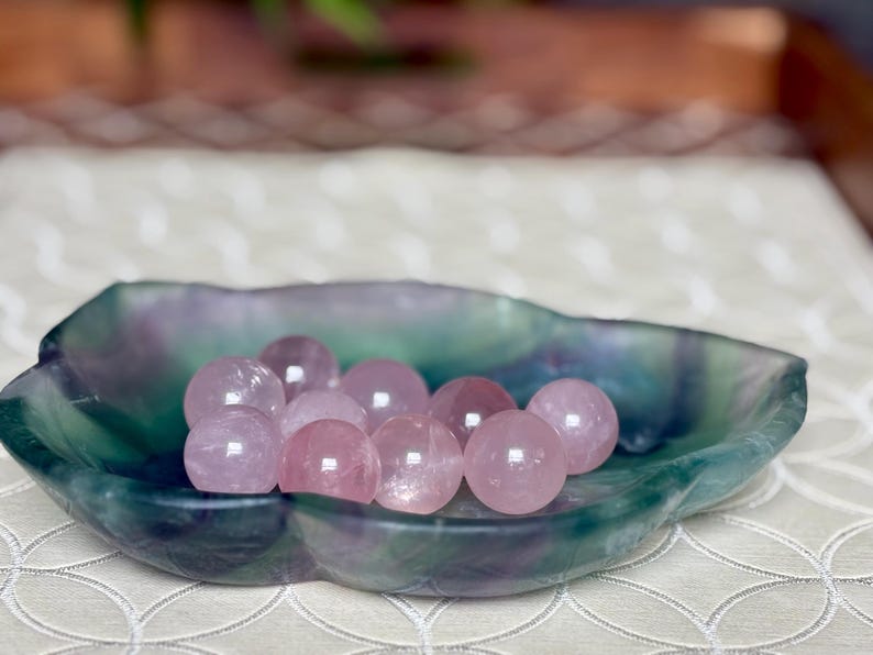 May include: A collection of pink rose quartz spheres displayed in a leaf-shaped dish. The dish has a marbled appearance with shades of green, blue, and purple. The spheres are smooth and translucent, reflecting light.