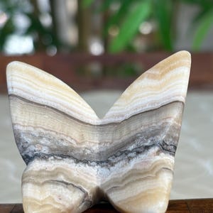 May include: A butterfly-shaped sculpture carved from layered stone. The stone features bands of cream, tan, gray, and black. The wings are spread wide, and the sculpture has a smooth, polished finish. The piece is a decorative item.