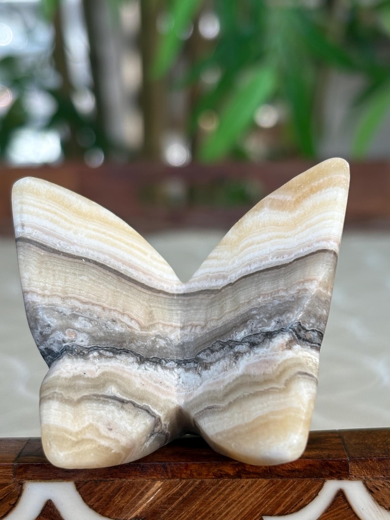 May include: A butterfly-shaped sculpture carved from layered stone. The stone features bands of cream, tan, gray, and black. The wings are spread wide, and the sculpture has a smooth, polished finish. The piece is a decorative item.