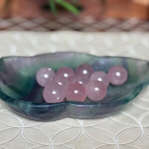 May include: A collection of pink rose quartz spheres displayed in a multi-colored fluorite dish. The dish is a mix of green, blue, and purple hues. The spheres are smooth and polished, reflecting light.