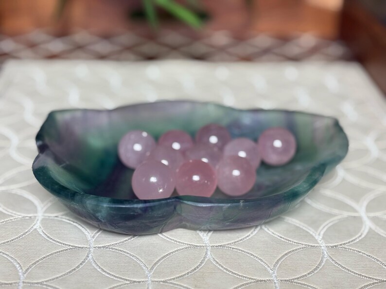 May include: A collection of pink rose quartz spheres displayed in a multi-colored fluorite dish. The dish is a mix of green, blue, and purple hues. The spheres are smooth and polished, reflecting light.