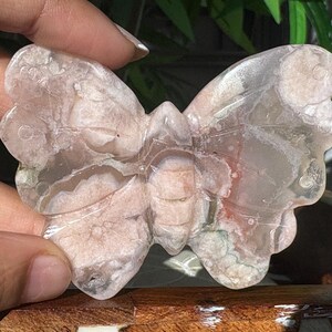 May include: A butterfly-shaped stone carving with a translucent appearance. The stone features a mix of pink, white, and clear colors, with visible patterns and textures. The butterfly is held by a hand, showcasing its detailed craftsmanship.