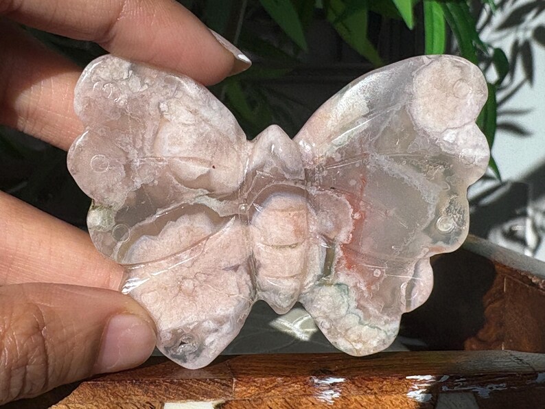 May include: A butterfly-shaped stone carving with a translucent appearance. The stone features a mix of pink, white, and clear colors, with visible patterns and textures. The butterfly is held by a hand, showcasing its detailed craftsmanship.