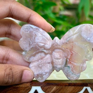 May include: A carved butterfly figurine in shades of pink, white, and tan. The wings have a detailed, textured appearance. The butterfly is held in a hand, showcasing its intricate design and natural stone appearance.