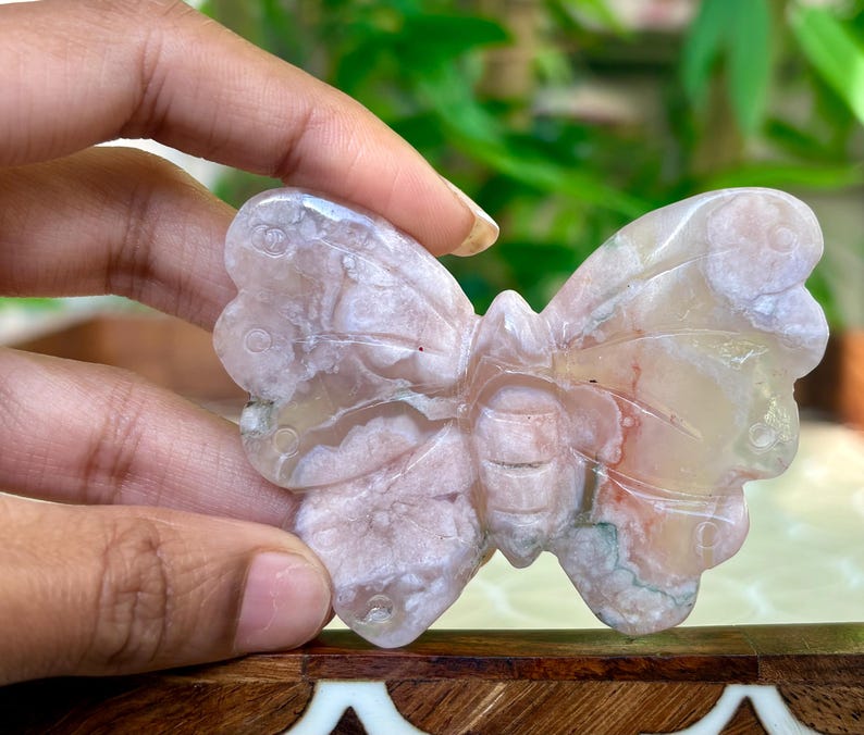 May include: A carved butterfly figurine in shades of pink, white, and tan. The wings have a detailed, textured appearance. The butterfly is held in a hand, showcasing its intricate design and natural stone appearance.