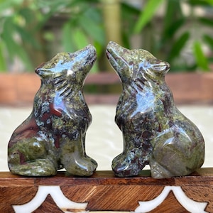 May include: Two carved stone wolf figurines in shades of green and burgundy. The wolves are in a seated position, heads tilted upwards. The figurines are displayed on a wooden surface with a decorative inlay.