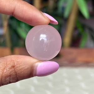 May include: A smooth, translucent rose quartz sphere. The sphere is a soft pink color and appears to be held between two fingers. The background is blurred, with hints of green foliage.