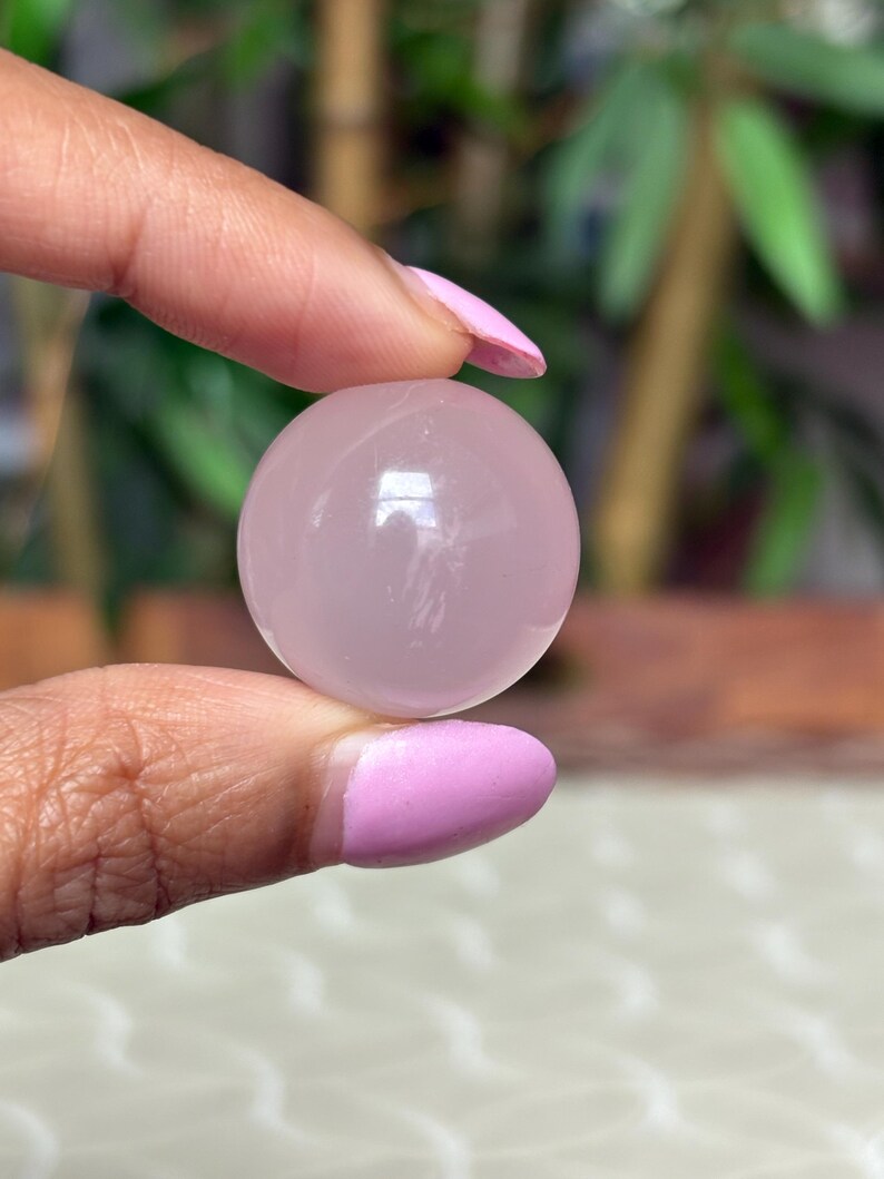 May include: A smooth, translucent rose quartz sphere. The sphere is a soft pink color and appears to be held between two fingers. The background is blurred, with hints of green foliage.