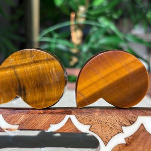 Puede incluir: Dos tallas pulidas de piedra ojo de tigre. Las piedras son de color marr&oacute;n c&aacute;lido con vetas doradas y tienen una forma curva &uacute;nica. Se exhiben sobre una superficie de madera decorativa.