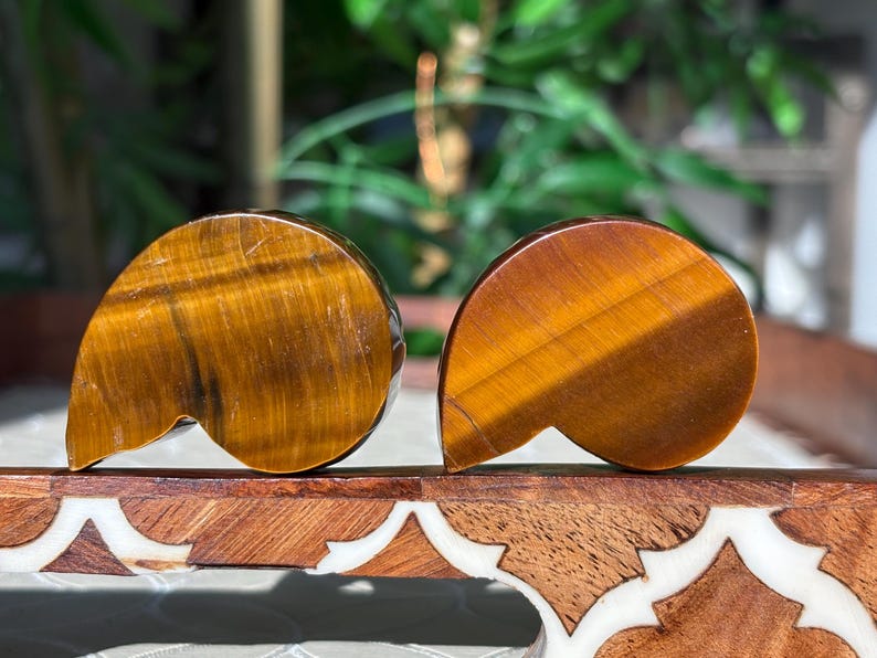 Puede incluir: Dos tallas pulidas de piedra ojo de tigre. Las piedras son de color marr&oacute;n c&aacute;lido con vetas doradas y tienen una forma curva &uacute;nica. Se exhiben sobre una superficie de madera decorativa.