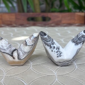 May include: Two V-shaped stone sculptures with unique patterns. One is a mix of beige, brown, and white, while the other is white with black and gray markings. The sculptures are displayed on a patterned surface.