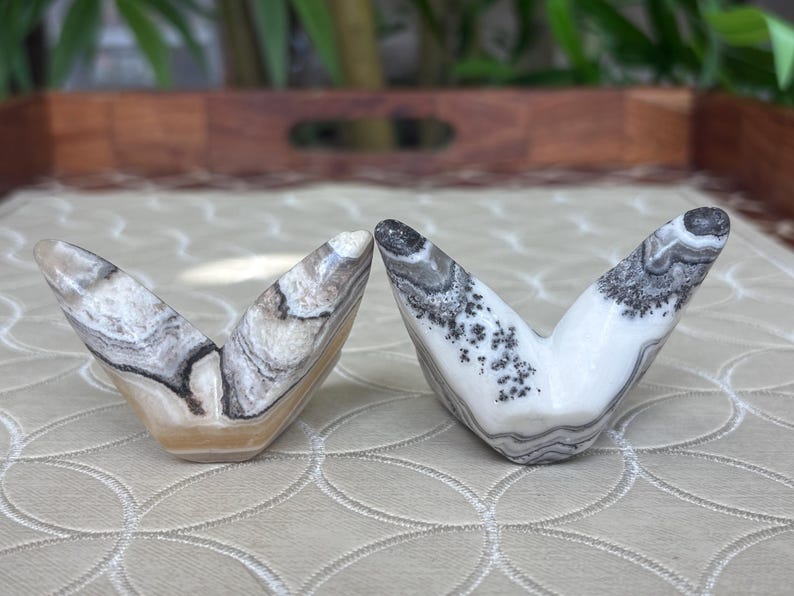May include: Two V-shaped stone sculptures with unique patterns. One is a mix of beige, brown, and white, while the other is white with black and gray markings. The sculptures are displayed on a patterned surface.