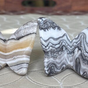 May include: Two butterfly sculptures carved from stone. One is primarily white and gray with a wavy pattern, the other has bands of tan, gray, and white. The wings are spread, and the bodies are touching.