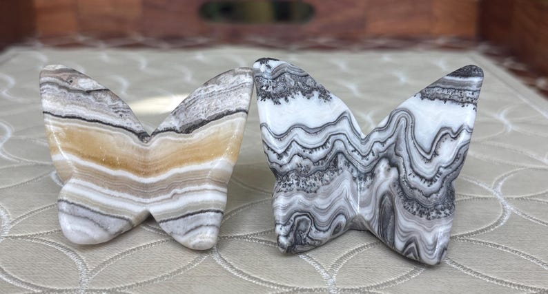 May include: Two butterfly sculptures carved from stone. One is primarily white and gray with a wavy pattern, the other has bands of tan, gray, and white. The wings are spread, and the bodies are touching.
