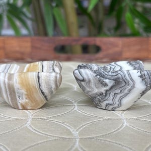 May include: Two decorative stone bowls with a unique shape. One bowl features a mix of beige, white, and brown tones, while the other has a black and white pattern. The bowls are placed on a patterned surface.