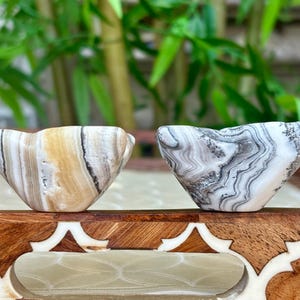 May include: Two small, carved stone bowls with unique patterns. One bowl has tan, brown, and white stripes, while the other features a gray and white marbled design. The bowls are displayed on a decorative wooden tray.