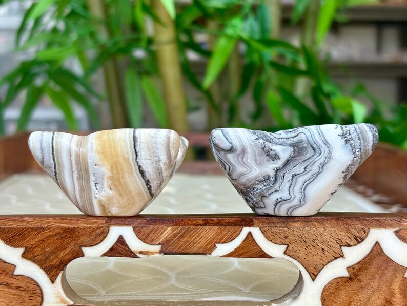 May include: Two small, carved stone bowls with unique patterns. One bowl has tan, brown, and white stripes, while the other features a gray and white marbled design. The bowls are displayed on a decorative wooden tray.