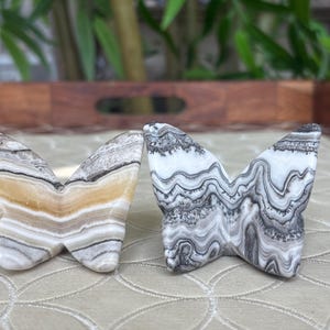 May include: Two butterfly-shaped stone sculptures. One is a mix of white, tan, and brown, while the other is primarily black and white. The wings have a layered, patterned appearance, and the sculptures are set on a patterned surface.