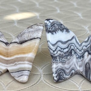 May include: Two butterfly-shaped stone sculptures. One is tan, brown, and white, the other is white, gray, and black. The wings have a layered, marbled appearance. These decorative items could be used as home decor.