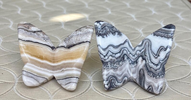 May include: Two butterfly-shaped stone sculptures. One is tan, brown, and white, the other is white, gray, and black. The wings have a layered, marbled appearance. These decorative items could be used as home decor.