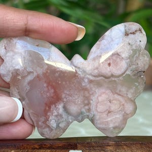 May include: A butterfly-shaped stone carving with a mix of pink, white, and brown hues. The natural stone has a polished surface and intricate patterns, showcasing the unique veining and color variations. The wings are detailed with rounded edges.