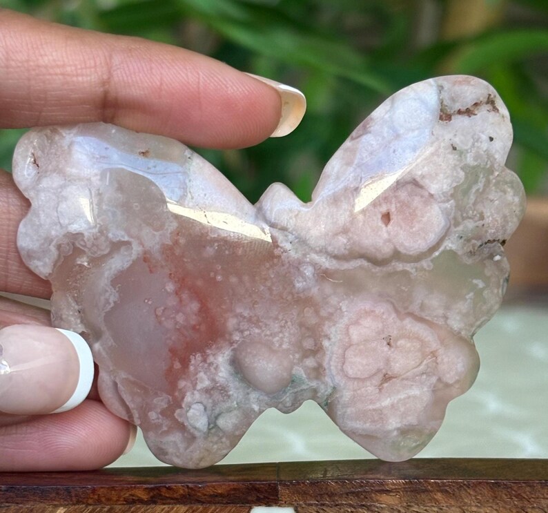 May include: A butterfly-shaped stone carving with a mix of pink, white, and brown hues. The natural stone has a polished surface and intricate patterns, showcasing the unique veining and color variations. The wings are detailed with rounded edges.