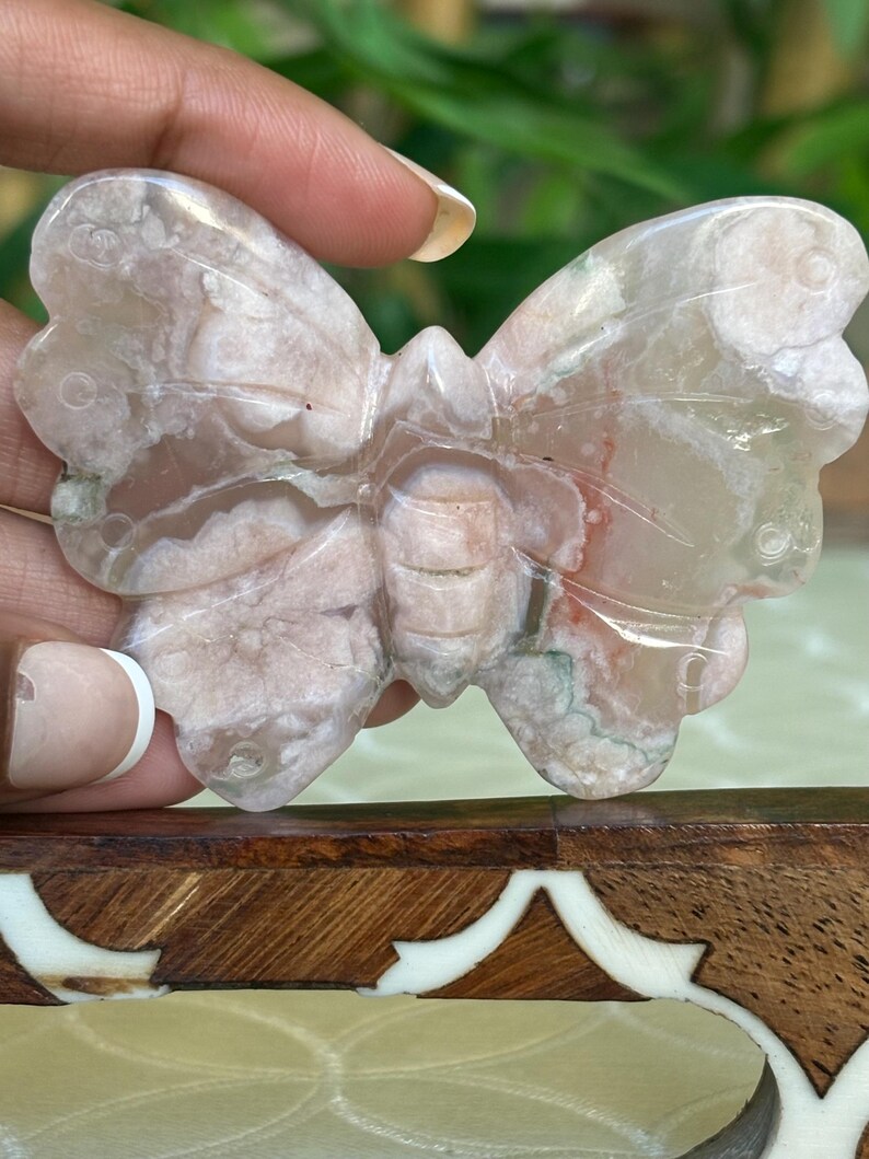 May include: A carved butterfly figurine in shades of pink, white, and gray. The wings have a detailed, veined pattern. The butterfly is held up against a blurred green background.