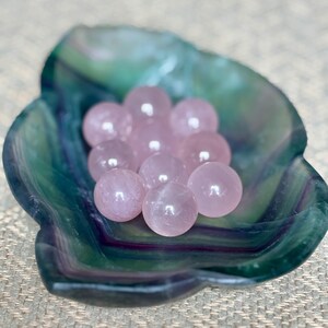 May include: A collection of small, polished rose quartz spheres nestled in a green and blue fluorite bowl. The spheres are a soft pink color and appear smooth. The bowl has a natural, organic shape and a marbled appearance.