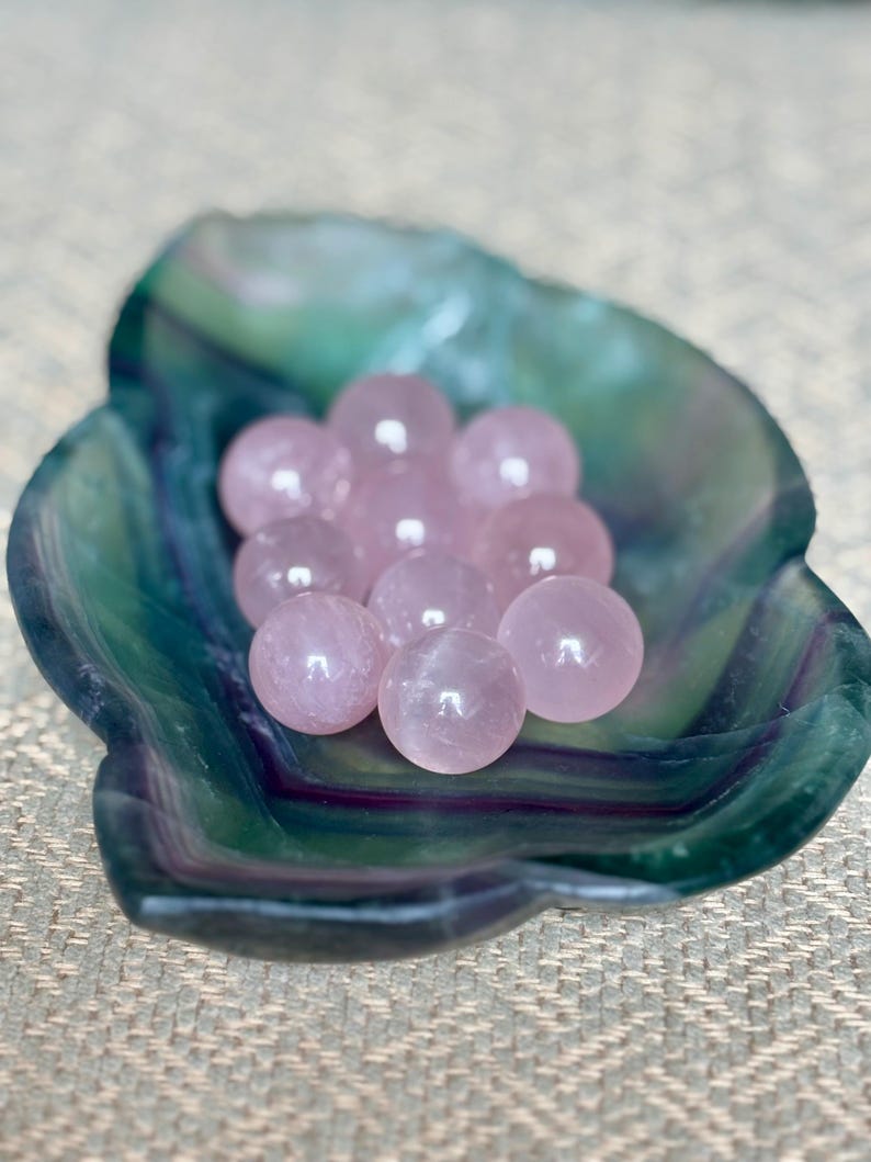 May include: A collection of small, polished rose quartz spheres nestled in a green and blue fluorite bowl. The spheres are a soft pink color and appear smooth. The bowl has a natural, organic shape and a marbled appearance.