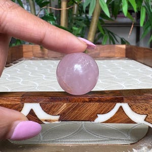 May include: A polished, spherical rose quartz crystal. The sphere is a soft pink color and appears smooth. The crystal is held in front of a decorative wooden tray with an inlaid design. The background features green foliage.