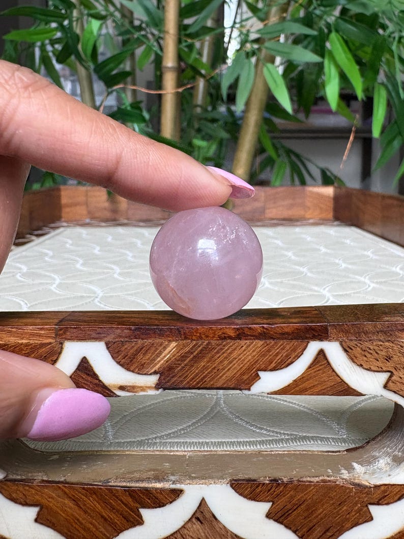 May include: A polished, spherical rose quartz crystal. The sphere is a soft pink color and appears smooth. The crystal is held in front of a decorative wooden tray with an inlaid design. The background features green foliage.