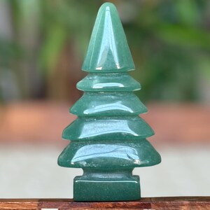 May include: A green gemstone Christmas tree figurine. The tree has a layered design with a pointed top and a square base. The gemstone has a translucent quality, and the tree is set against a blurred background.