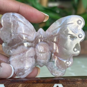 May include: A carved butterfly figurine made of translucent, light pink and white stone. The butterfly has detailed wing patterns and circular accents. The figurine is held by a hand, showcasing the intricate details and natural variations in the stone.